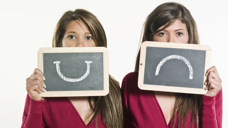 Zwei Frauen, eine mit einem lachenden, eine mit einem traurigen Smiley auf einem Schild.