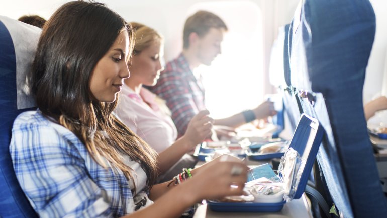 Zwei junge Frauen und ein junger Mann, die im Flugzeug nebeneinander sitzen, nehmen eine Bordmahlzeit zu sich.
