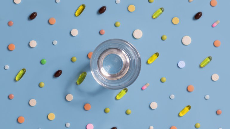 Auf einem blauen Untergrund steht ein Glas Wasser, drumherum liegen viele bunte Pillen, Dragées, Kapseln und Tabletten.