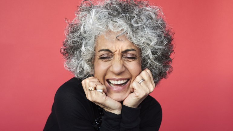 Eine lachende Frau mit lockigen grauen Haaren vor rotem Hintergrund
