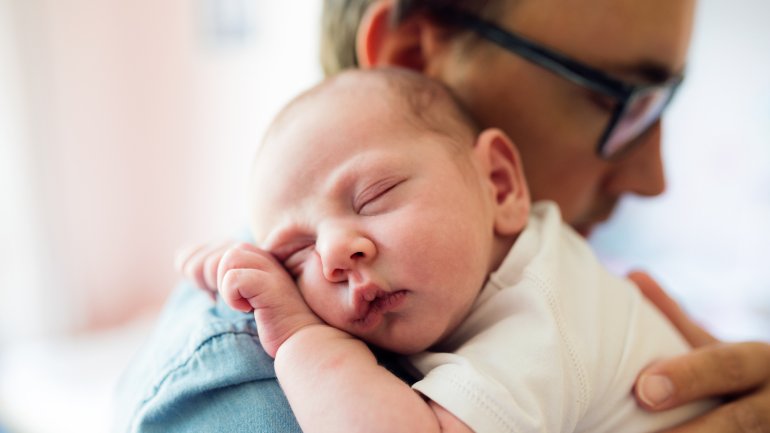 Ein junger Mann mit Brille trägt ein schlafendes Baby.