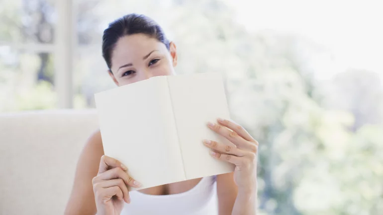 Eine junge lächelnde Frau hält ein Buch vors Gesicht, sodass ihr Mund verdeckt ist.